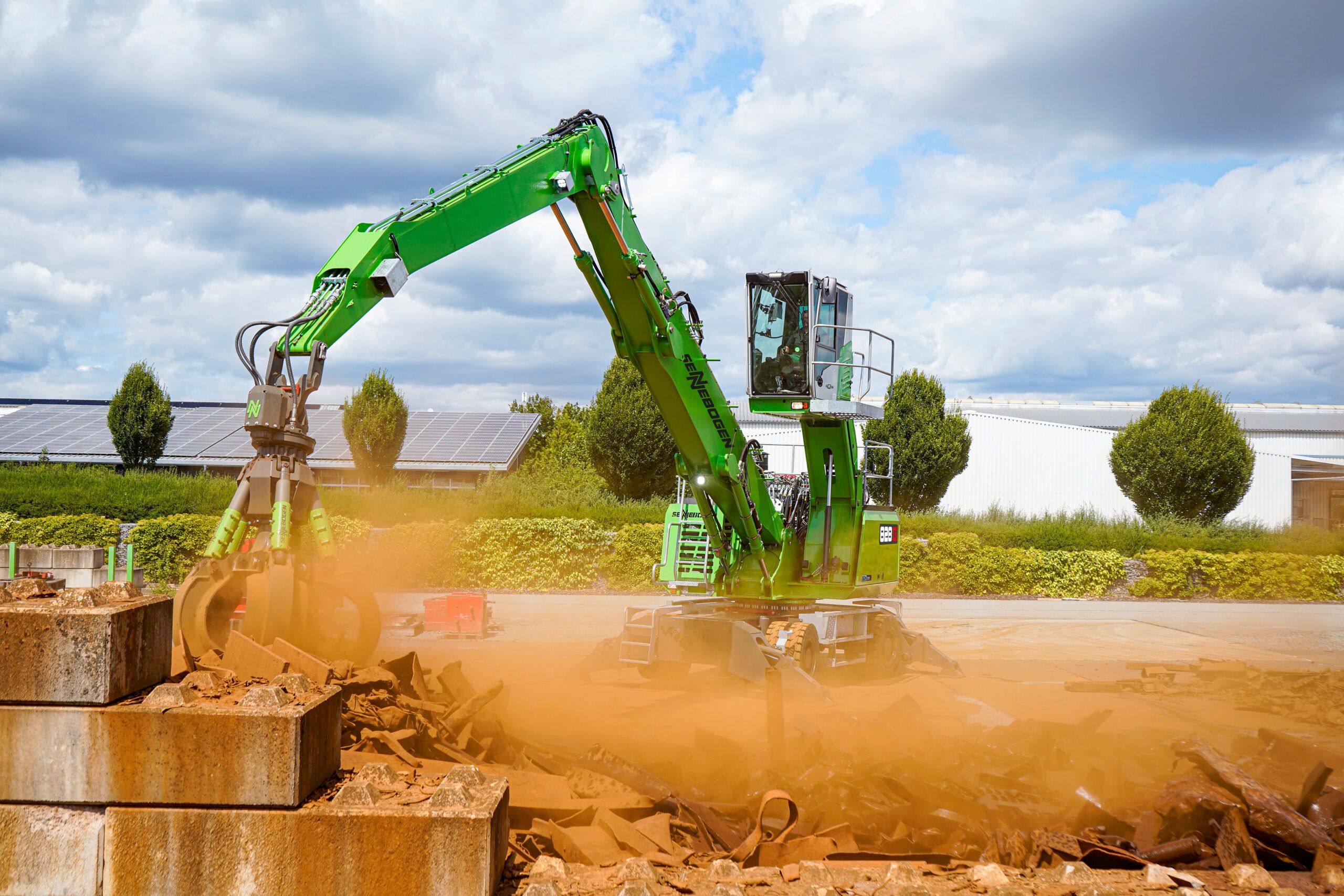 A green Sennebogen 828 G material handler on a wheeled undercarriage working a pile of scrap beside large concrete blocks and kicking up dust, with a solar-panelled building roof and trees in the background.