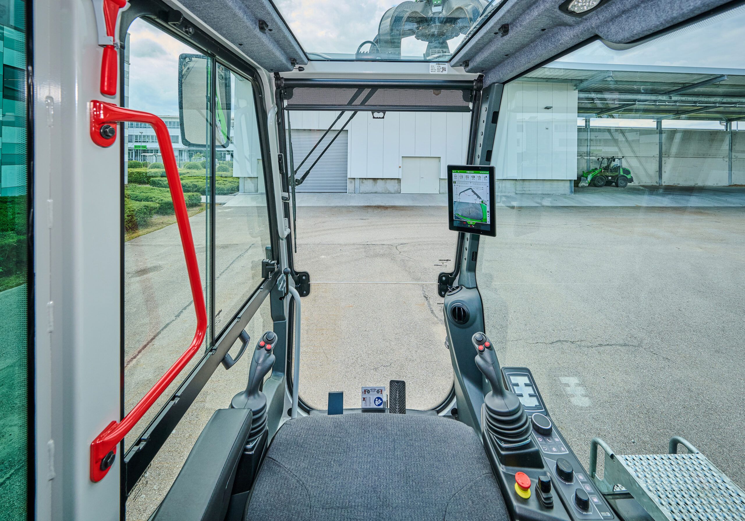 The view from the operator’s seat inside the 828 G Maxcab: two joysticks, a 10in touchscreen mounted on the right-hand pillar showing the SENcon display, a red grab handle on the glazed left-hand door, and a paved yard visible through the large windows.