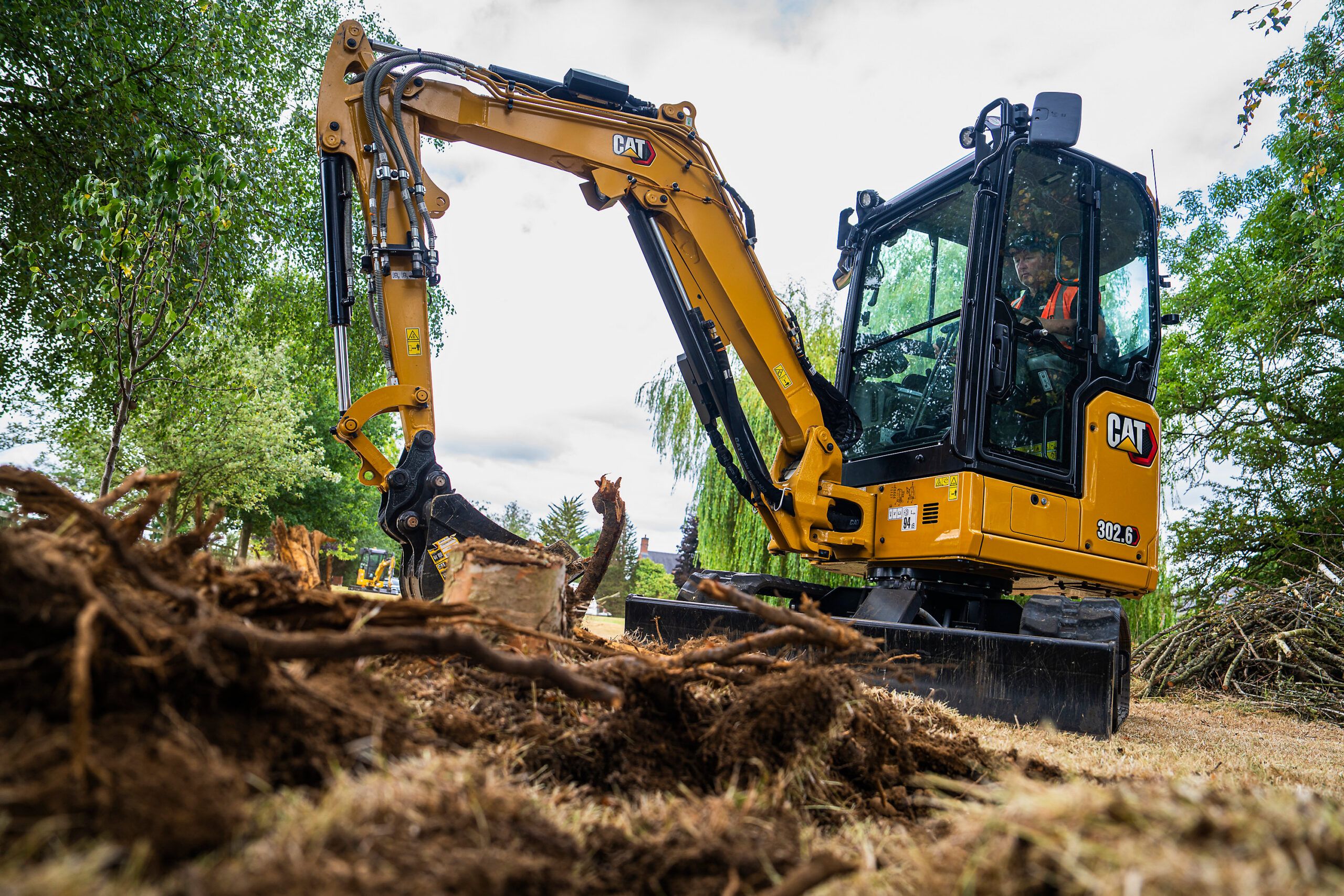 : A yellow Cat 302.6 mini excavator positioned beside uprooted tree roots and brash on grassland, with an operator in the cab.