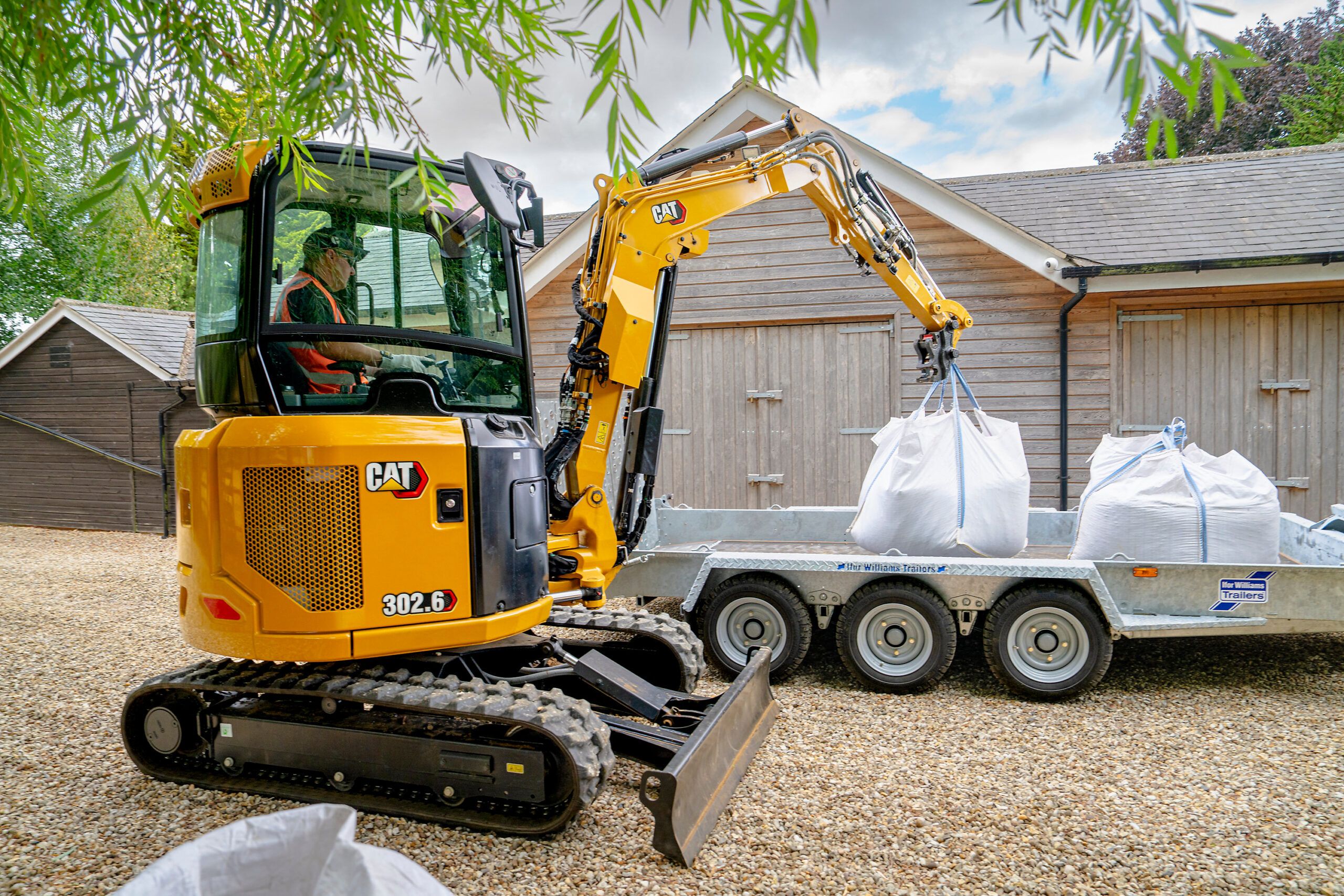 A yellow Cat 302.6 mini excavator lifting a large white bulk bag onto a flatbed trailer in a gravel yard.