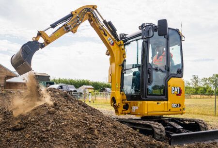Caterpillar adds two models to Next Generation mini excavator range A yellow Cat 302.4 CR mini excavator with an operator in orange hi-vis digging into a mound of soil on a rural site.