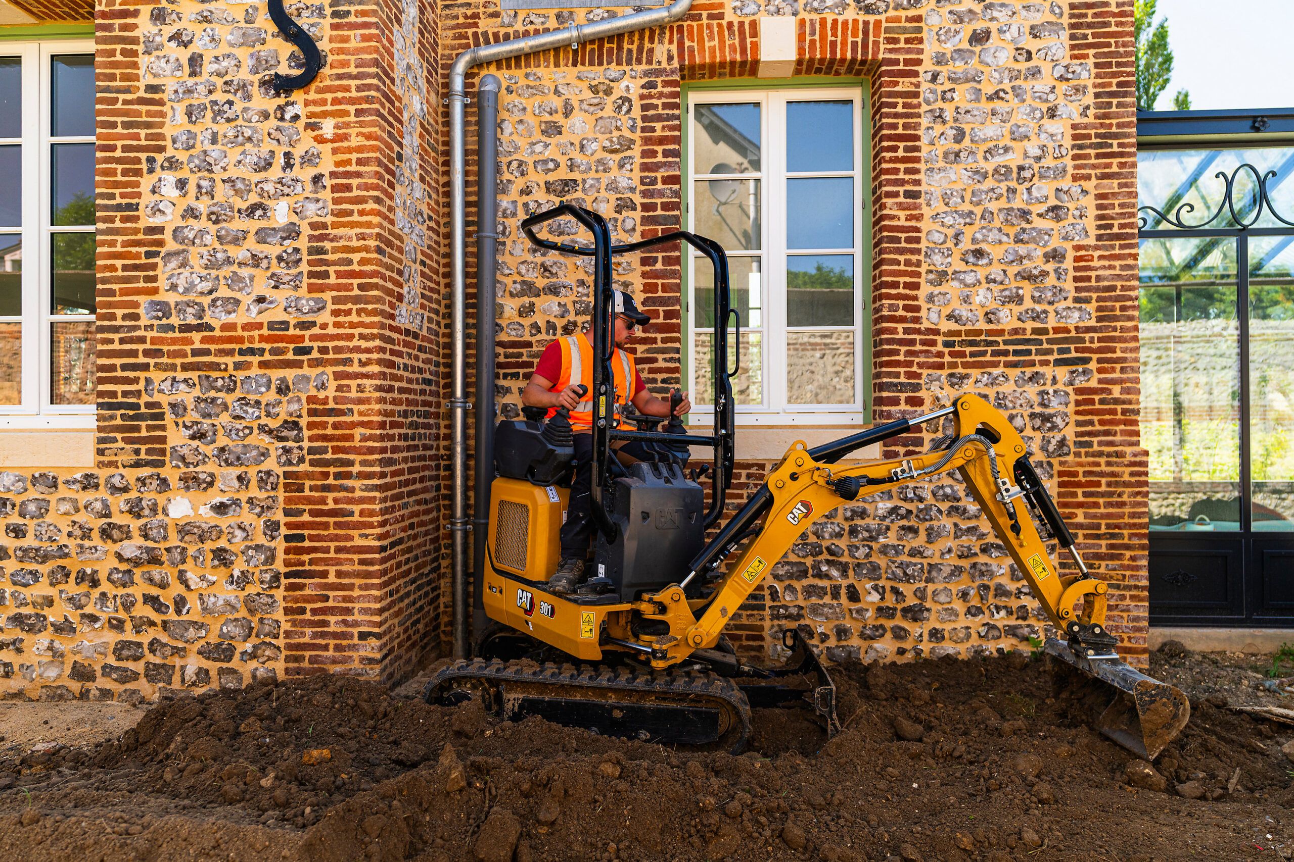 A yellow Cat 301 CR micro excavator digging a soil trench hard against the corner of a brick and flint building.
