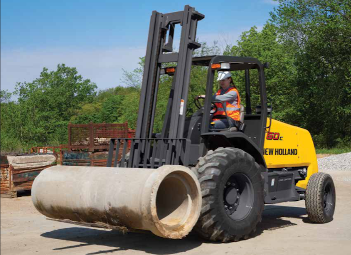 A yellow New Holland F60C rough terrain forklift carrying a large concrete pipe on its forks, driven by an operator in a hi-vis vest in an outdoor materials yard.
