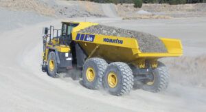 : A yellow Komatsu HM460-6 articulated dump truck loaded with aggregate, driving around a curved dirt haul road in a quarry and throwing up dust.