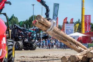 Alt text: A hydraulic crane grapple lifts several debarked logs at an outdoor forestry machinery demonstration, with exhibitor banners and spectators in the background.
