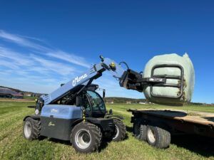 : A pale-blue Mona Nova electric telehandler lifting a wrapped round bale onto a flatbed trailer in a grass field, with farm buildings behind.