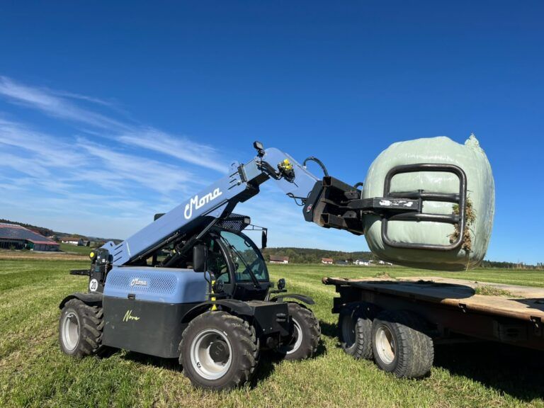 Mona Nova to debut electric telehandler at EuroTier : A pale-blue Mona Nova electric telehandler lifting a wrapped round bale onto a flatbed trailer in a grass field, with farm buildings behind.