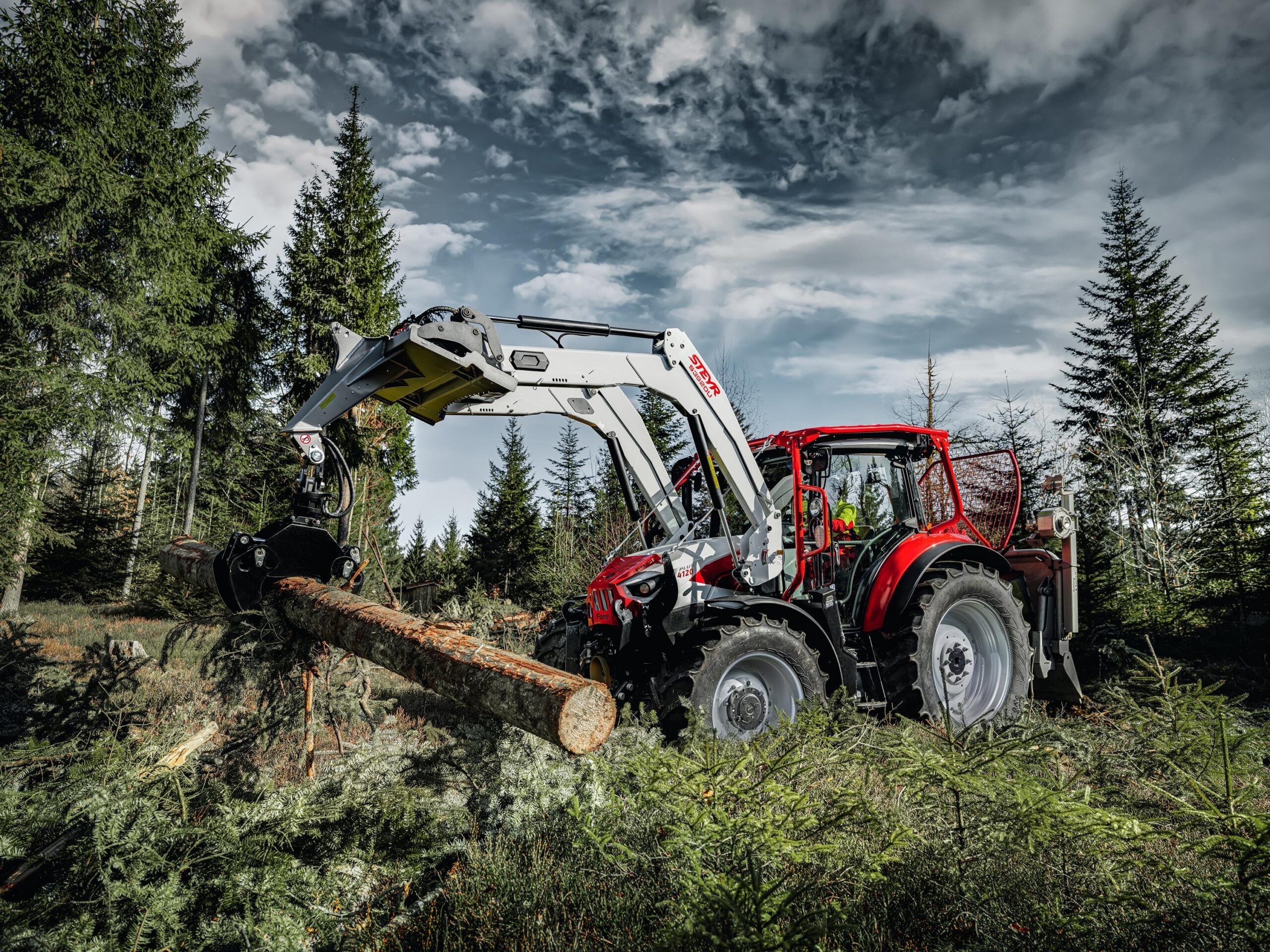A red Steyr Plus tractor in forestry specification, fitted with a front loader and grapple lifting a felled log in a spruce forest under a dramatic cloudy sky.