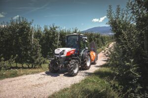 A white and red Steyr Spezial 4120F specialty tractor on a gravel track between rows of apple trees, towing a mounted sprayer, with mountains in the background.