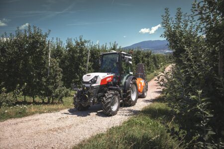 A white and red Steyr Spezial 4120F specialty tractor on a gravel track between rows of apple trees, towing a mounted sprayer, with mountains in the background.