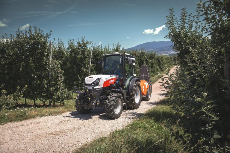 Steyr launches Spezial F CVT tractor for orchard growers A white and red Steyr Spezial 4120F specialty tractor on a gravel track between rows of apple trees, towing a mounted sprayer, with mountains in the background.
