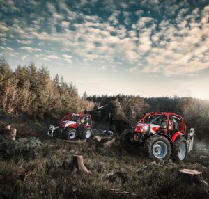 Two red Steyr forestry tractors in a cleared woodland at golden hour, one carrying a rear-mounted timber crane, among tree stumps and felled logs