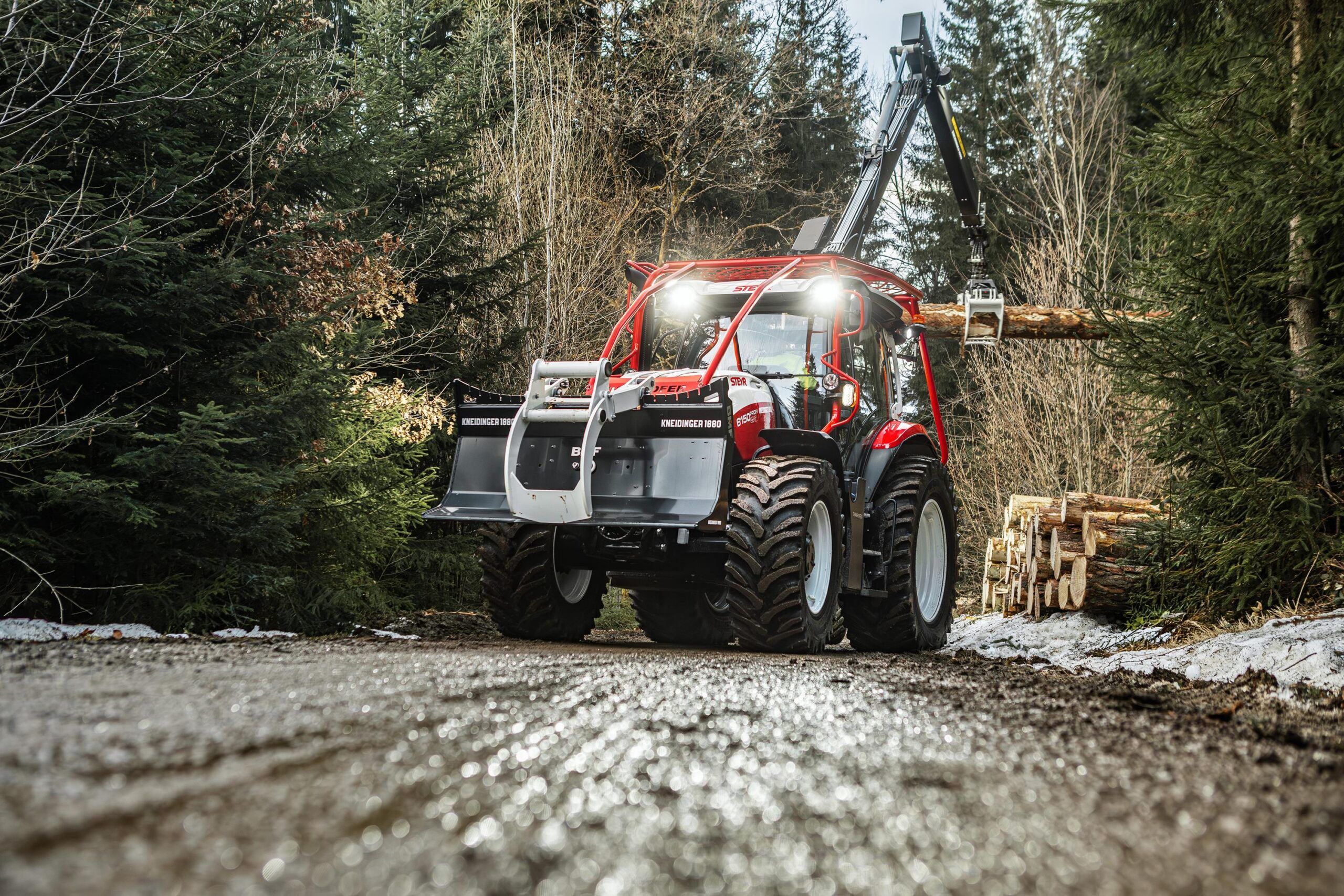 A red Steyr Profi CVT forestry tractor with a protective cab frame and roof lights on a wet forest track in winter, with stacked timber alongside.