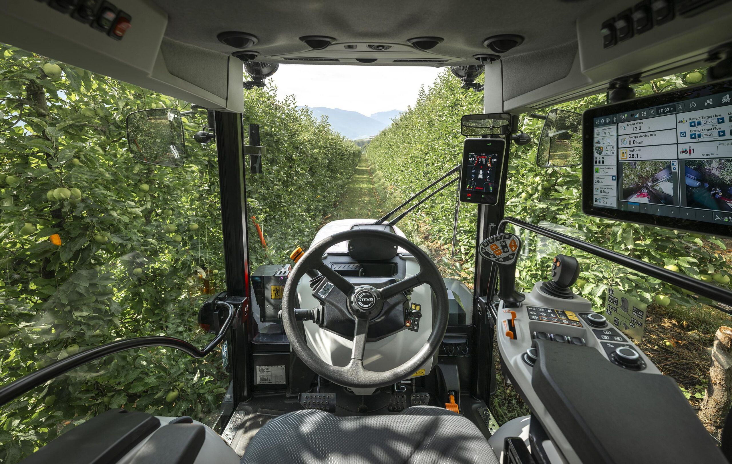 Cab interior of a Steyr Spezial F CVT tractor driving between rows of apple trees, showing the steering wheel, joystick armrest and two display screens