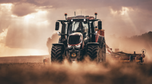 Front view of a Valtra sixth-generation T Series tractor working in a dusty field at dusk, towing a trailed implement, with an operator in the cab.
