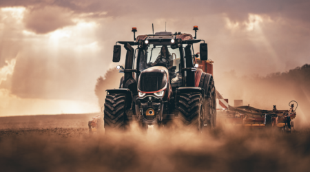 Front view of a Valtra sixth-generation T Series tractor working in a dusty field at dusk, towing a trailed implement, with an operator in the cab.