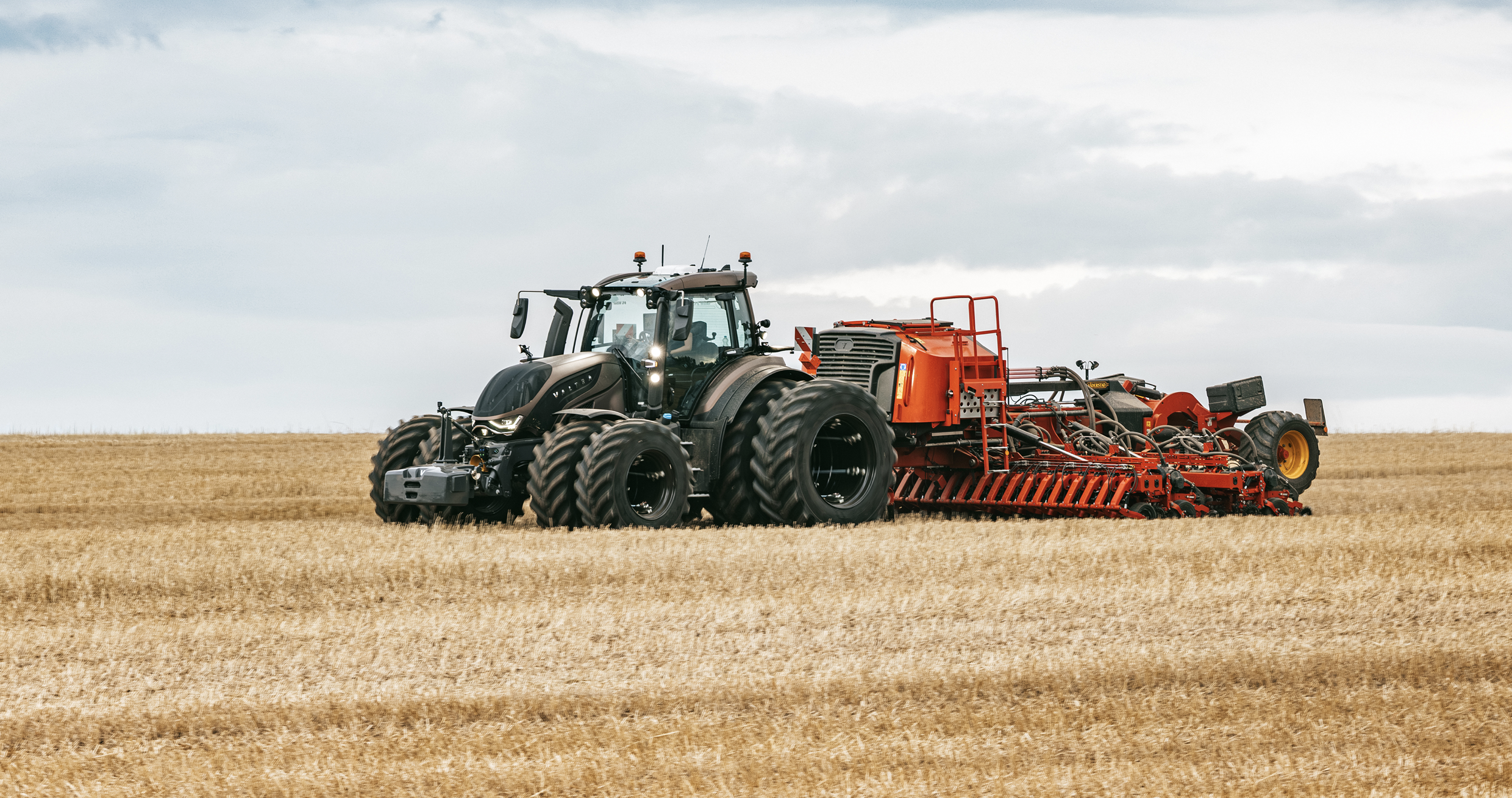 Side view of a Valtra sixth-generation T Series tractor on dual wheels towing a red seed drill across a harvested field.