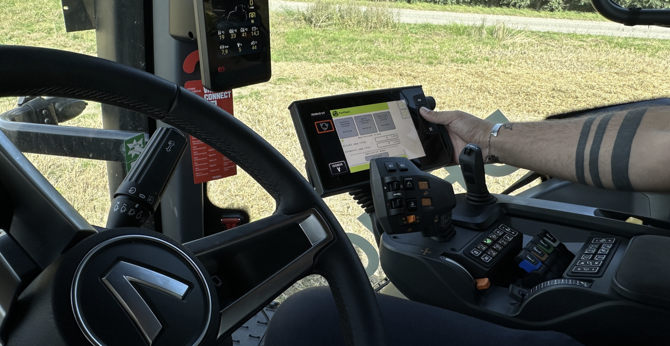 Inside a Valtra tractor cab, an operator’s hand touches the ISOBUS display showing the PerPlant crop-sensor interface, beside the SmartTouch armrest controls and steering wheel.