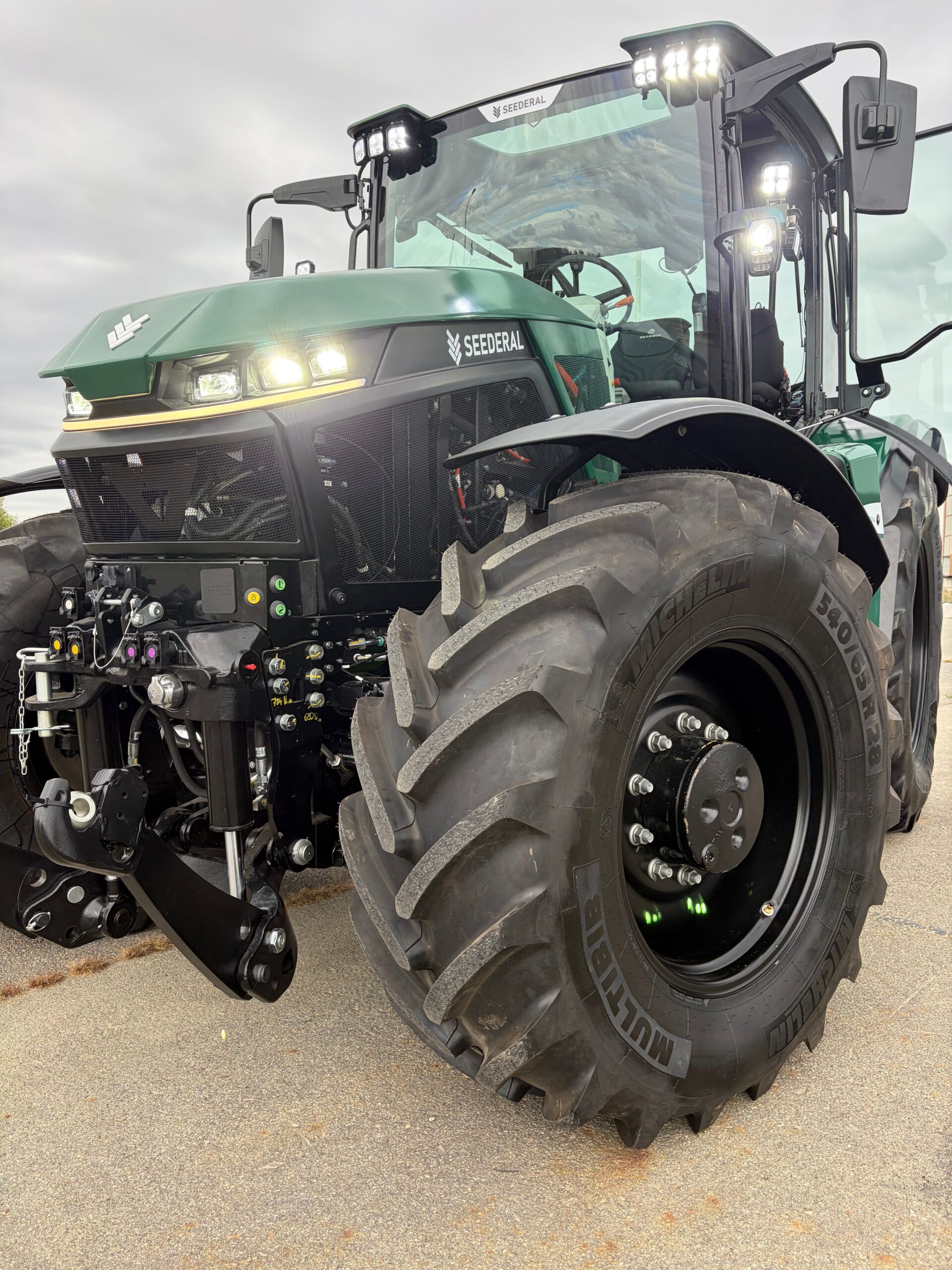 Low-angle close-up of the front of a green Seederal electric tractor with its LED work lights illuminated, showing the front linkage and a large agricultural tyre.
