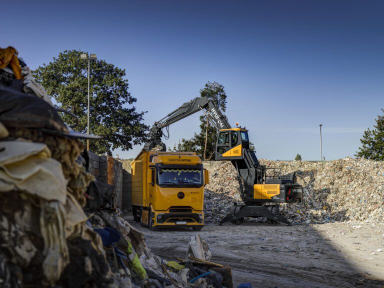 Hyundai launches 28-tonne HW270A MH material handler A yellow and black Hyundai HW270A MH wheeled materials handler, fitted with an orange-peel grapple and with its cab raised, loads waste into the open body of a yellow container truck at an outdoor recycling yard under a clear sky.