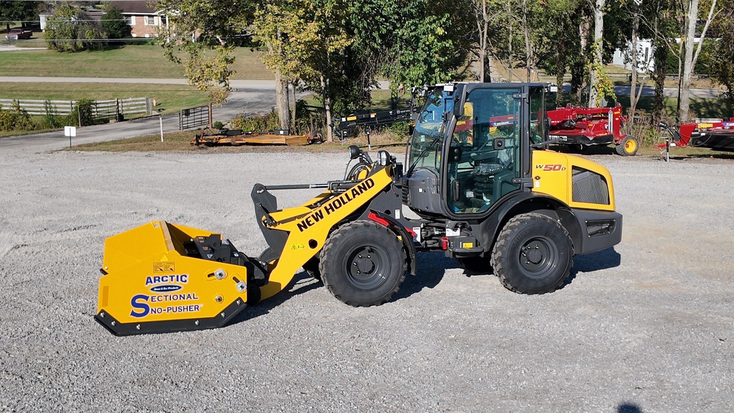 : A yellow New Holland W50D compact wheel loader fitted with a snow-pusher attachment, parked on a gravel lot.