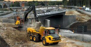 A Volvo crawler excavator loads excavated material into a Volvo articulated hauler at a bridge construction site, with roadworks and a concrete bridge structure behind.