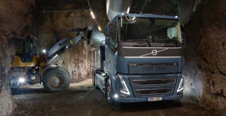 A dark blue Volvo FH Electric truck with a tipping trailer stands in a rock-walled underground tunnel, while a yellow Volvo wheel loader lifts material in the background.