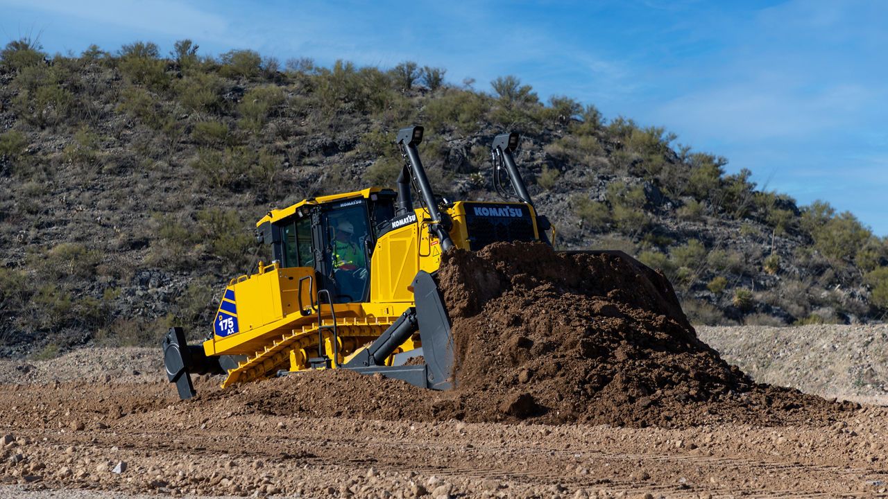 A yellow Komatsu D175AX-10 bulldozer with an operator in the cab, pushing a large pile of earth on a dirt site with a scrub-covered hillside behind.