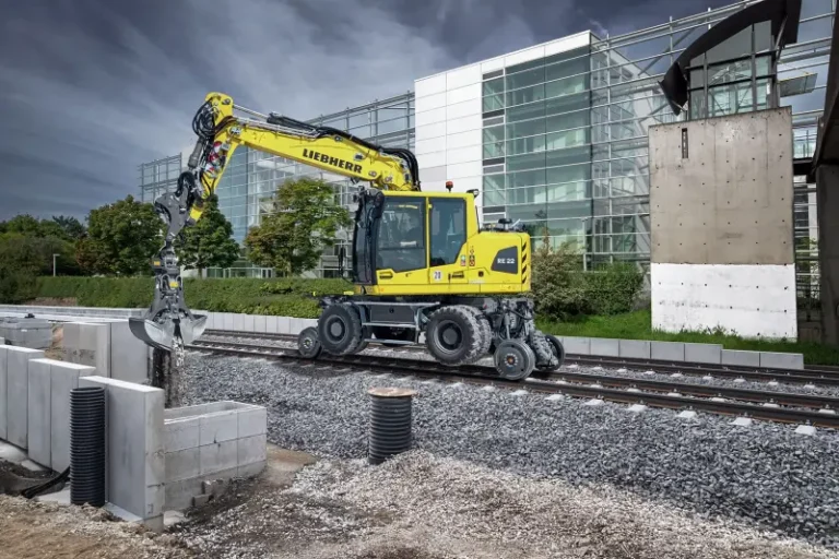 Liebherr unveiled RE 22 M Litronic rail excavator at InnoTrans A yellow Liebherr RE 22 M railroad excavator fitted with a grab attachment working on a railway track, its road-rail undercarriage resting on the rails, with a modern glass building behind